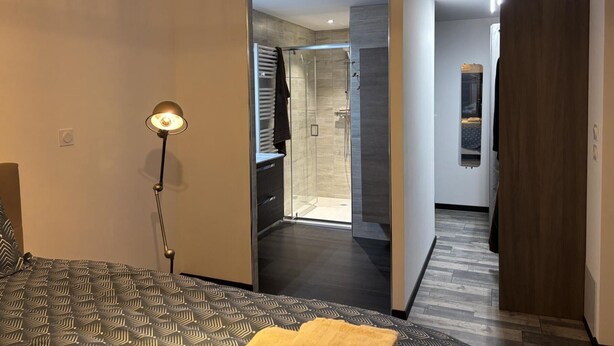 A modern hotel room with a bed, a bedside lamp, a bathroom with a shower, and a hallway leading to another room.