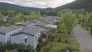 A row of single-story cabins with flat roofs, surrounded by greenery and hills in the background. - Mobile home for 6 ppl with terrace (Saint-Maurice-d'Ibie)