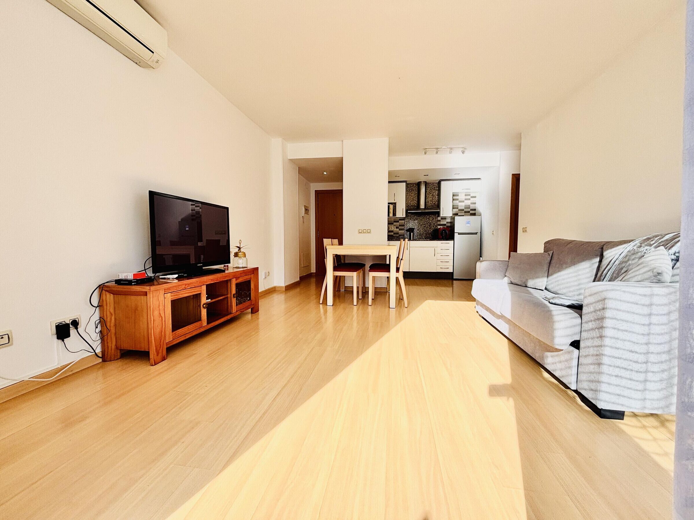 Een moderne woonkamer met een flatscreen televisie, een houten entertainmentkast en een grijze bank. Er is een eethoek met een tafel en stoelen, en een keuken op de achtergrond.
