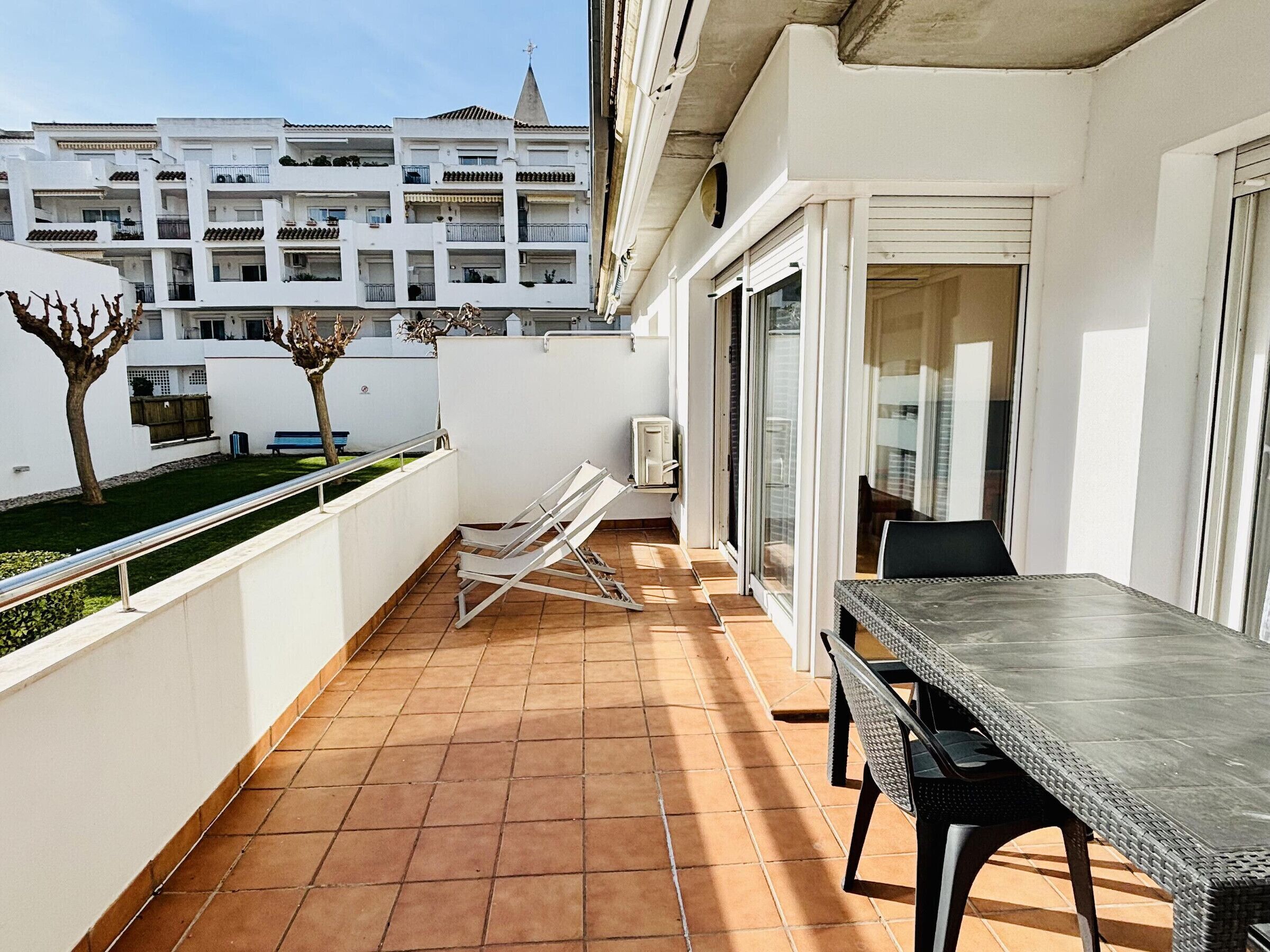 A balcony with a table and chairs, a small garden, and a view of a building with multiple balconies.