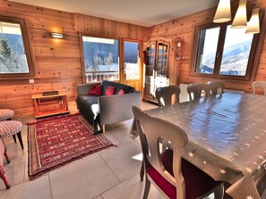 Ein gemütliches Blockhaus-Interieur mit einer Couch, einem Esstisch und Stühlen, mit Holzwänden und großen Fenstern mit Blick auf einen verschneiten Berg.