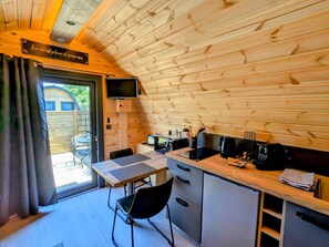 A compact kitchen with wooden walls, a dining table, and a TV mounted on the wall. - Scandinavian wooden pod for 2 people with WiFi (Rainville)