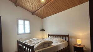 Un dormitori amb sostre de fusta, un llit amb roba de llit blanca, una tauleta de nit amb una làmpada i una finestra amb cortines.