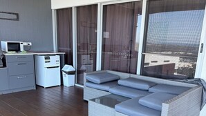 Un salon moderne avec un canapé d’angle gris, une petite kitchenette et une grande porte-fenêtre coulissante donnant sur un espace extérieur.