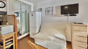 Kompakti oleskelutila, jossa on pieni keittiönurkkaus, valkoinen sohva, televisio ja puinen kaappi.