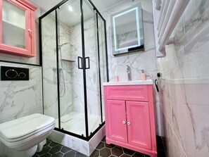 Un baño con ducha de vidrio, inodoro blanco, muebles rosa y salpicadero de mármol.