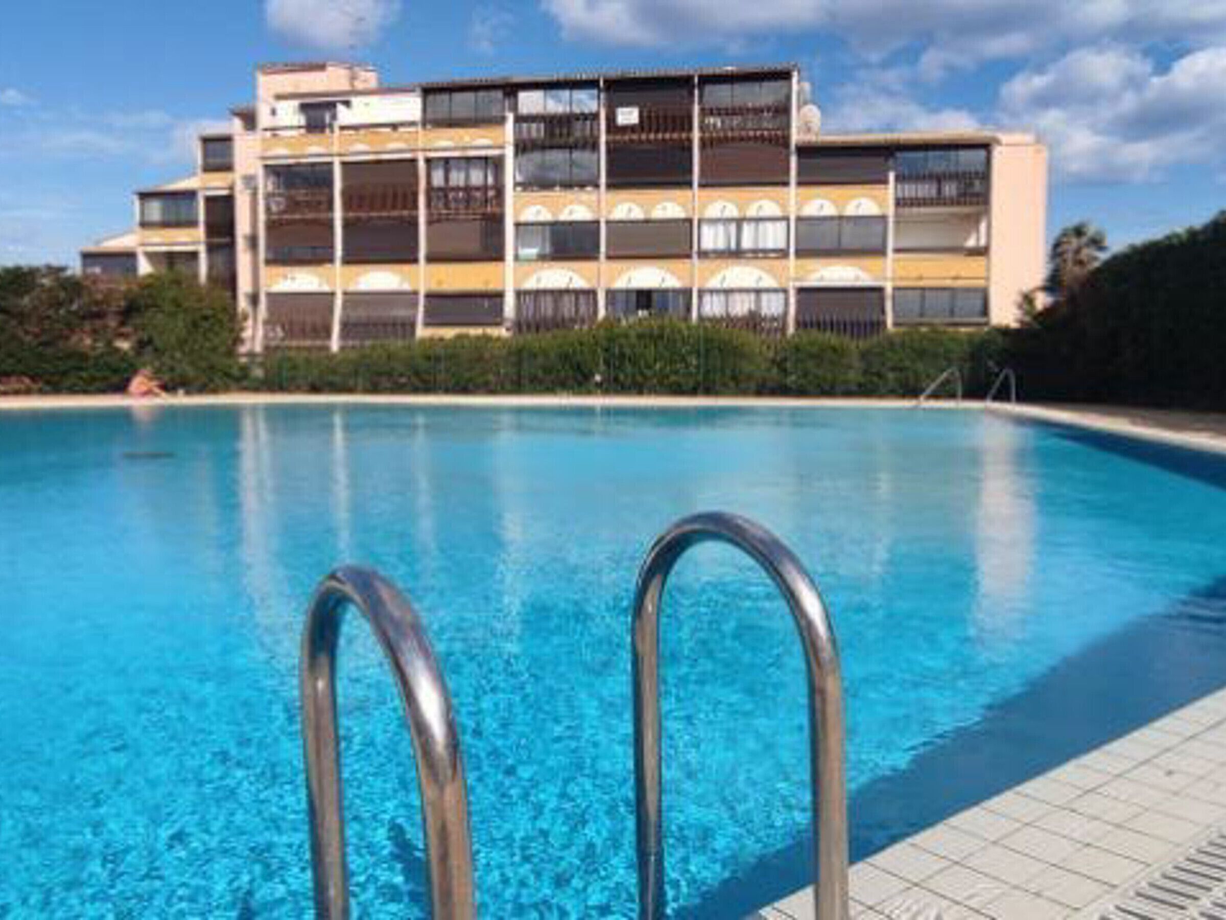 Una piscina con escaleras metálicas en primer plano y un edificio de varios pisos con balcones al fondo.