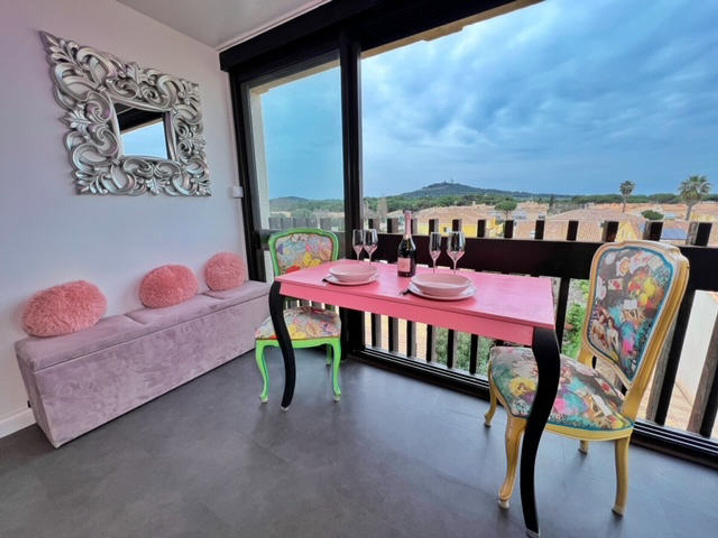 Un balcón con una mesa rosa dispuesta para dos personas, un banco acolchado y sillas con tapicería floral.