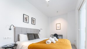 Un dormitorio moderno con un cabecero tapizado gris, ropa de cama blanca, un edredón color mostaza y una pequeña mesa redonda con un control remoto.