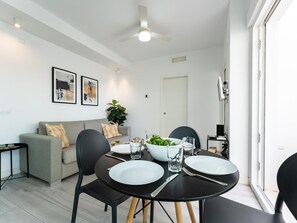 Ein moderner Essbereich mit einem runden Tisch, schwarzen Stühlen und einem grauen Sofa. An der Wand befinden sich zwei gerahmte Kunstwerke, ein Deckenventilator und eine Tür zu einem anderen Raum.