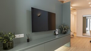 Una habitación moderna con una televisión de pantalla plana en la pared, una pequeña mesa con un jarrón de flores y un espejo que refleja la habitación.