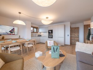 Eine moderne Küche mit weißen Schränken, ein Essbereich mit Holztisch und -stühlen sowie ein Wohnzimmer mit Sofa und Couchtisch.
