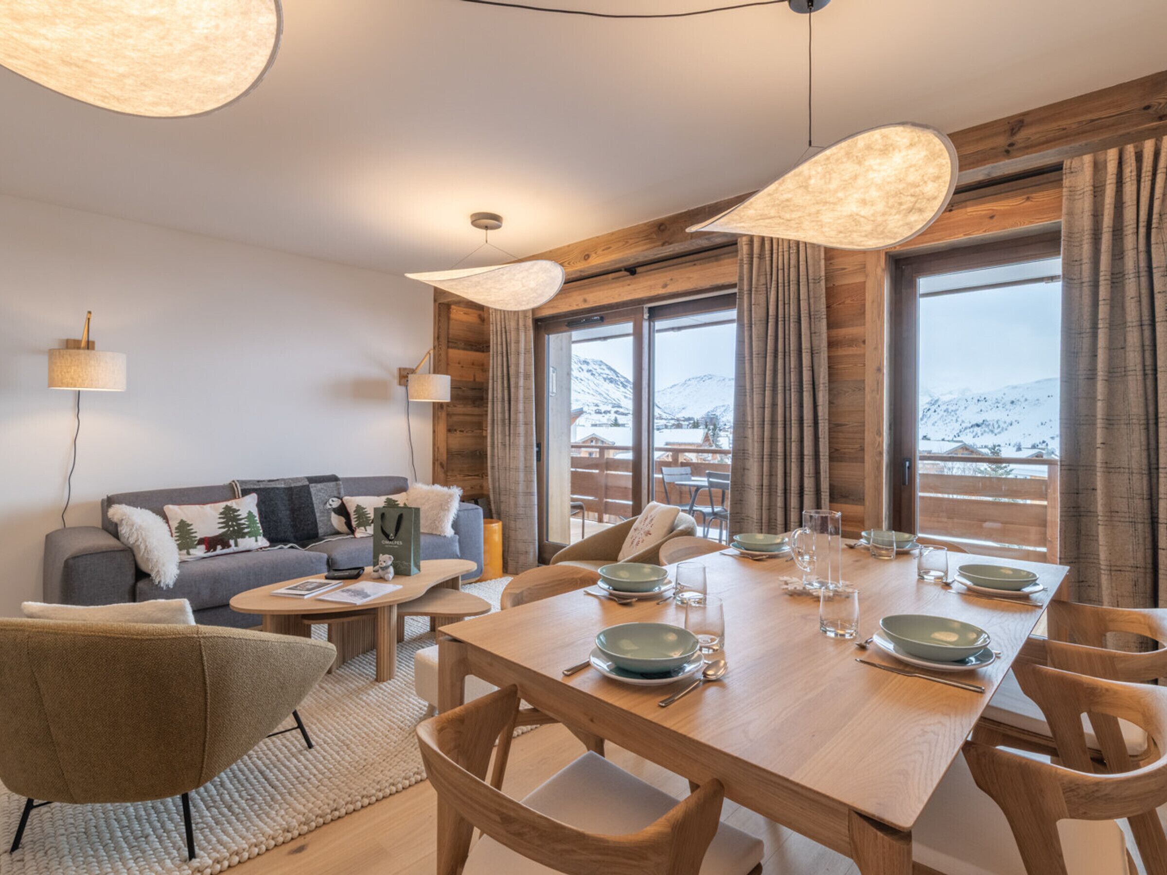 A dining area with a wooden table set for four, surrounded by wooden chairs. There is a sofa with cushions and a lamp in the background.