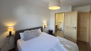 Un dormitorio con cama, lámparas de noche, un armario de madera y un baño a la vista a través de una puerta abierta.