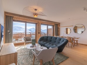 A modern hotel room with a large window offering a mountain view, a cozy seating area with a dark armchair and two chairs, a wooden dining table, and a unique ceiling light fixture. - Elegant apartment, 6 pax, ski-in/ski-out in Huez (Huez)