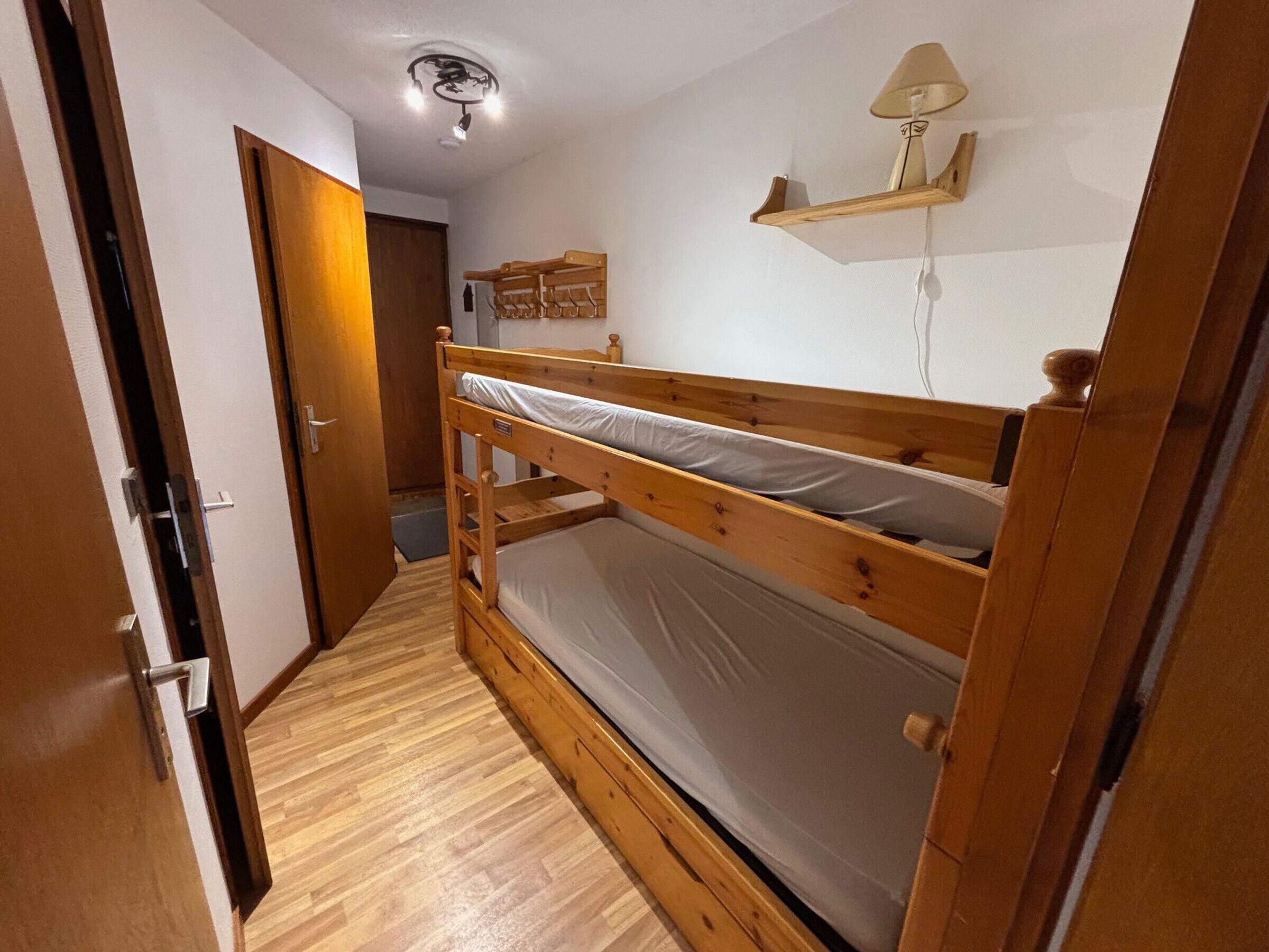 A bunk bed room with a wooden floor, a ceiling light, a wooden door, and a wall-mounted shelf with a lamp.