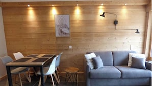 A room with a wooden wall, a gray sofa, a dining table with chairs, and a small round table with a lamp. - Renovated studio for 4 with balcony in Grand-Bornand (Le Grand-Bornand)