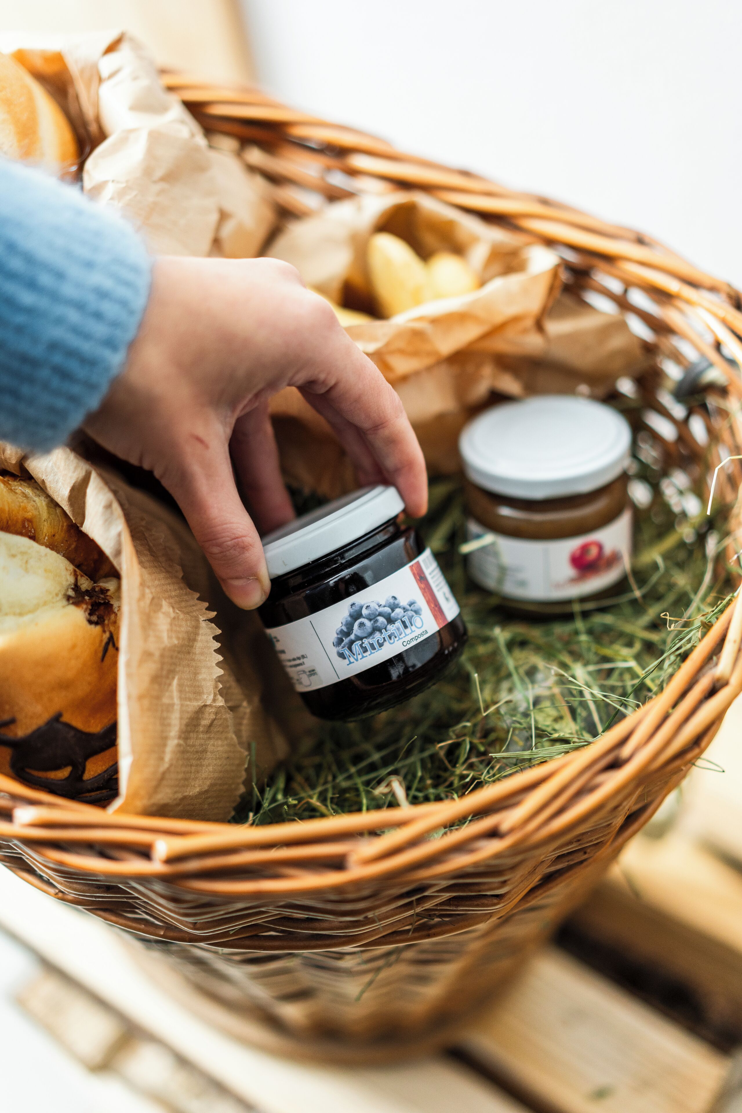 Eine Hand hält ein Marmeladenglas in einem Korb aus Weidengeflecht, der mit Brot und einem weiteren Marmeladenglas gefüllt ist.
