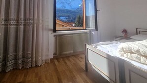A bedroom with a wooden floor, a bed, a window with a mountain view, and a radiator. - Appartment le Gemme Dolomites escape (Dosoledo)