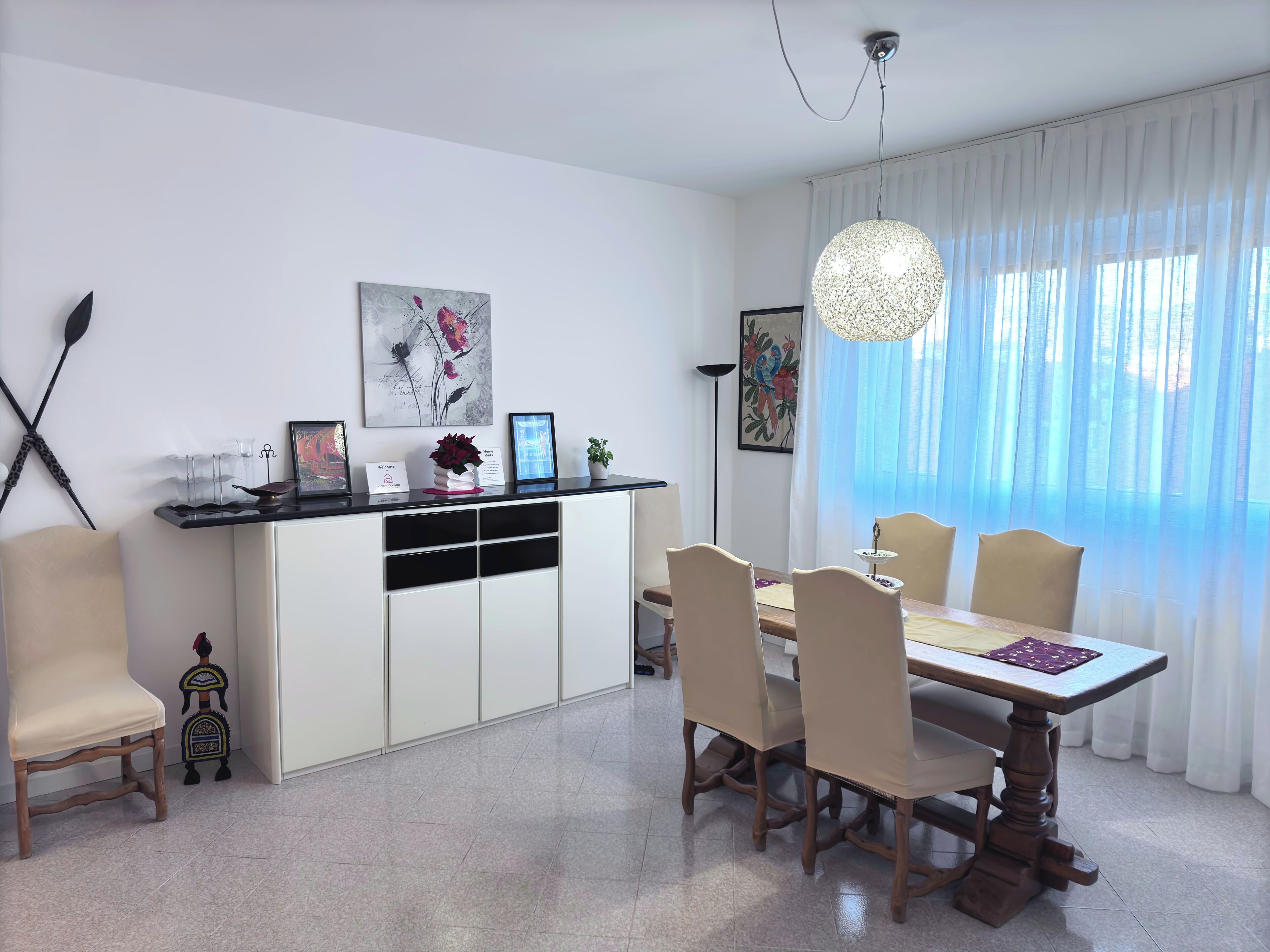 Un comedor con una mesa y sillas de madera, un armario blanco con utensilios de cocina y obras de arte en las paredes.