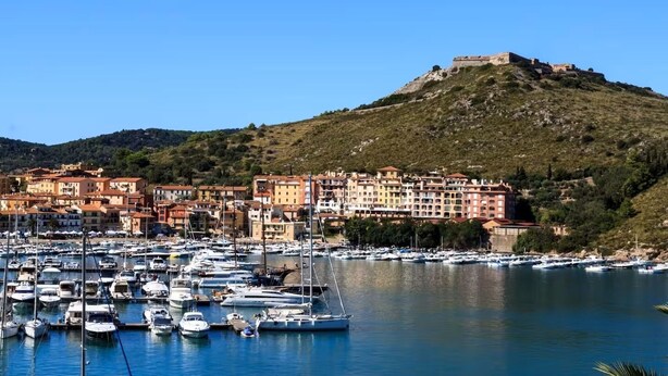 A coastal town with a historic fort on a hill, a marina with numerous boats, and colorful buildings along the waterfront. - Tenuta San Crescenzio (Toscana)