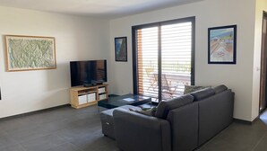 Ein Wohnzimmer mit grauem Sofa, einem Flachbildfernseher auf einem Holzboden und einem Glastisch. An der Wand hängen zwei gerahmte Bilder und es gibt ein großes Fenster mit Jalousien.