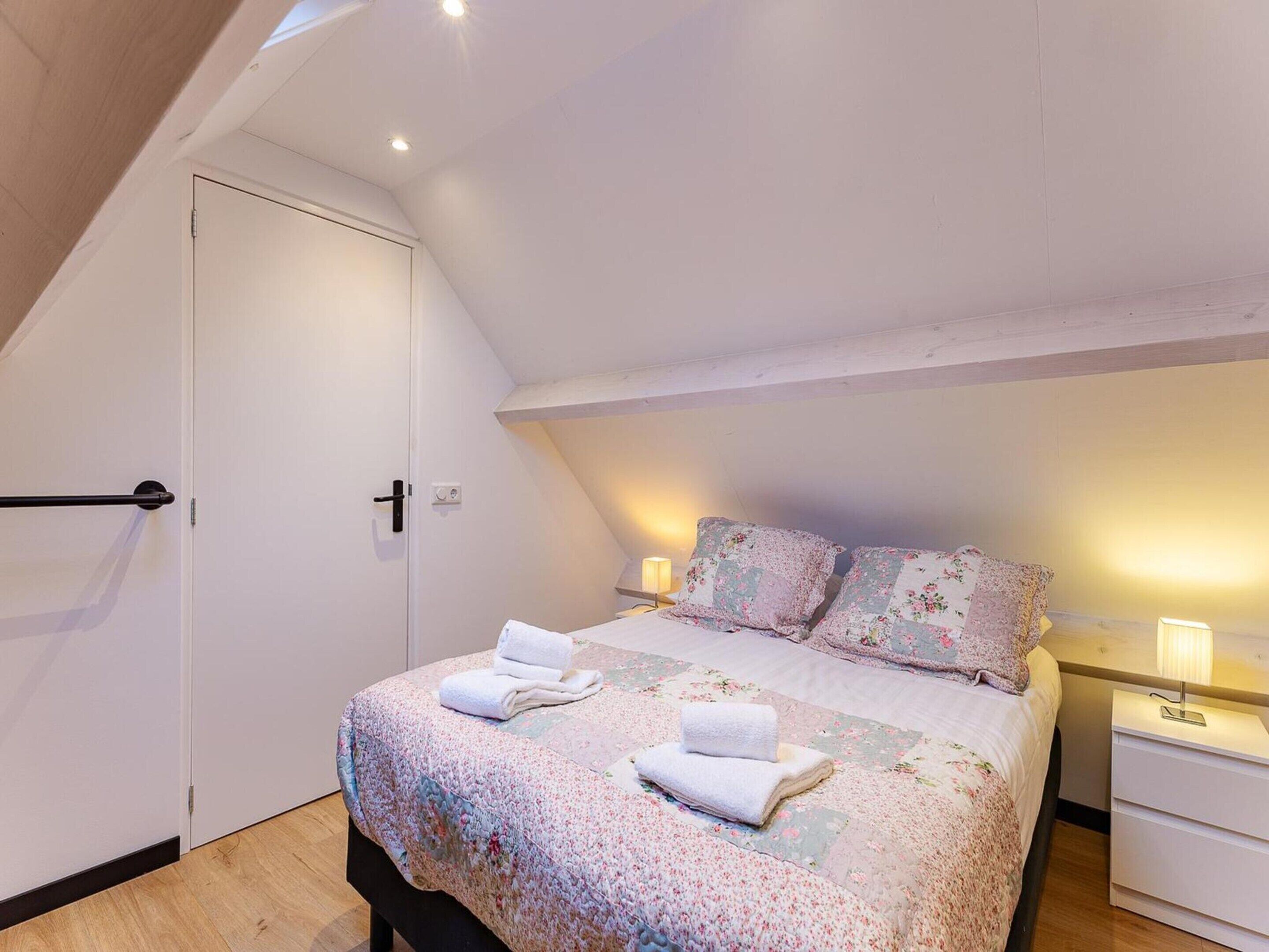 Een slaapkamer met een bed, kussens, een nachtkastje, een deur en een plafondlamp.