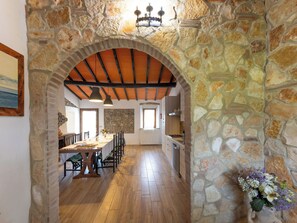 Un arco de piedra que da acceso a un comedor con suelo de madera y una cocina al fondo.