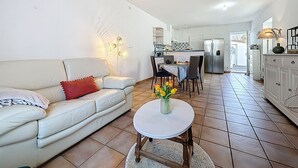 A living room with a white leather sofa, a small round table with a vase of flowers, and a kitchen in the background. - Sanoubott by Interhome (La Bernerie-en Retz)