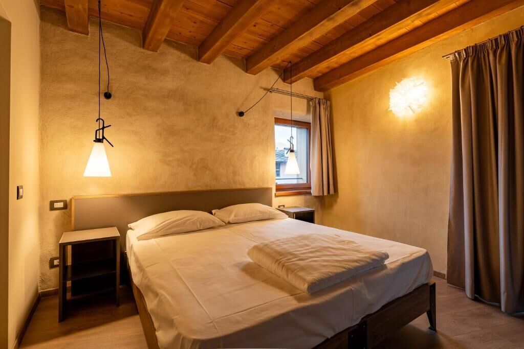 A bedroom with a wooden ceiling, a bed with white linens, a nightstand, a wall-mounted lamp, and a window with curtains.