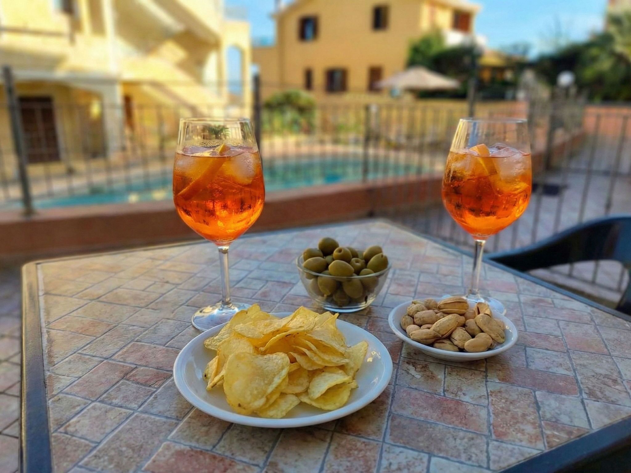 Two glasses of orange beverage on a table with plates of chips and nuts, a bowl of olives, and a pool in the background.