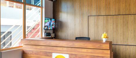 A reception desk with a wooden finish and a logo, a television screen, and a view of a staircase.
