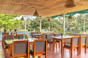 An outdoor dining area with tables and chairs, surrounded by lush greenery and a thatched roof. - Itsy Hotels Sands & Serenity (Gokarna)