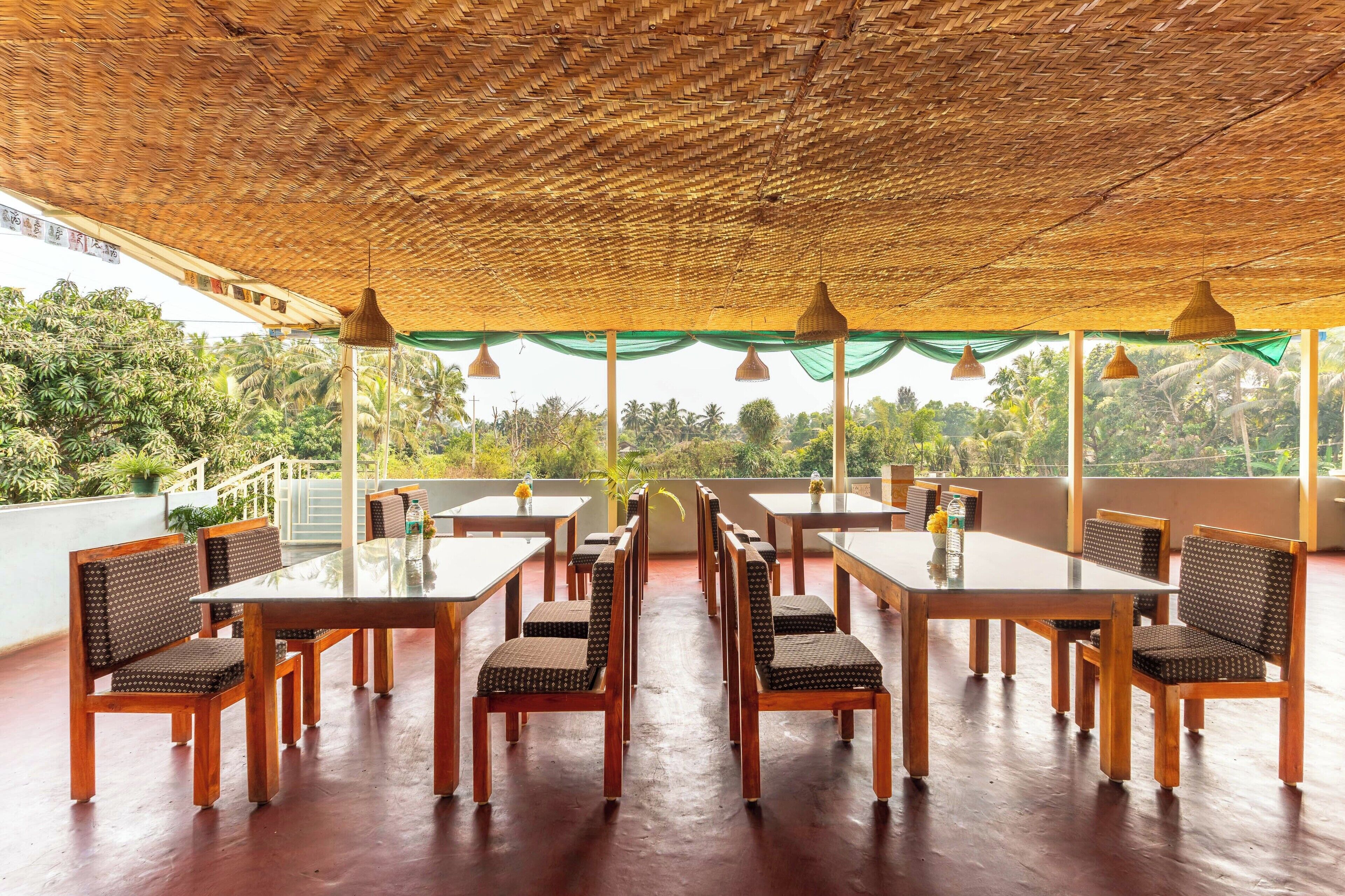 A covered outdoor dining area with tables and chairs, a thatched roof, and a view of greenery.