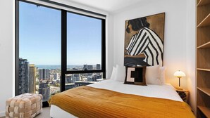 A modern bedroom with a large window, a city view, a bed with a patterned throw, a side table with a lamp, and a wooden bench. - Queensbridge Luxe Living Next to Crown on the River. Pool • Gym • Sauna Steam (Southbank)