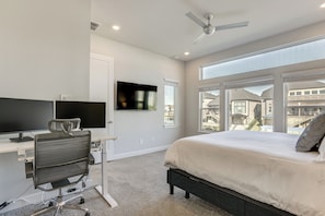 Un dormitorio con cama, escritorio con computadora, silla, televisor montado en la pared y ventanas amplias con vista a edificios.