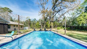 A rectangular swimming pool with a blue tile border, surrounded by a grassy area with trees and a house in the background. - 5 Mi to Silver Springs State Pk: Gem w/ Game Room! (Ocala)