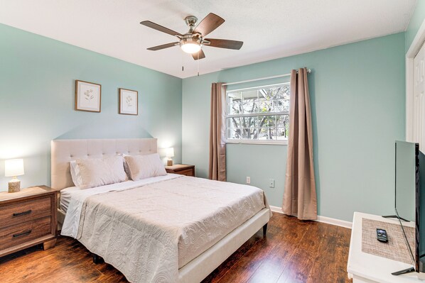 A bedroom with a bed, nightstands, a ceiling fan, a window with curtains, and a television on a stand.