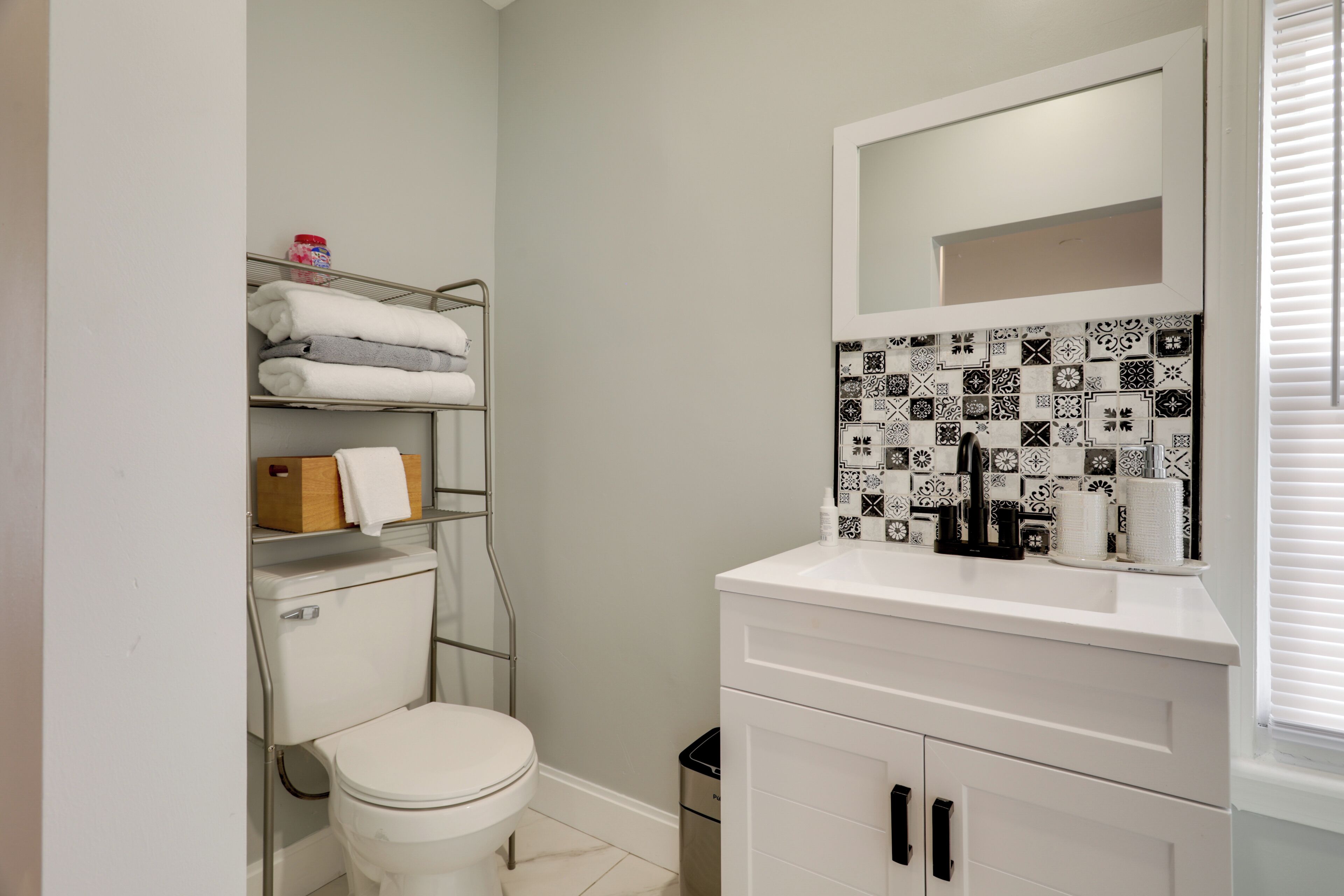A bathroom with a toilet, a sink with a mirror, a shelf with towels, and a patterned backsplash.