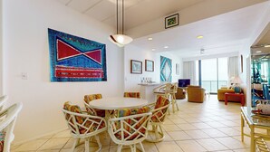 A spacious living room with a round dining table surrounded by wicker chairs, a large abstract painting on the wall, and a balcony with a view. - 2BR Penthouse Paradise w/ Front-row Gulf Views (South Padre Island)