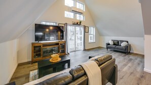 A spacious loft-style living room with a high ceiling, a large flat-screen TV, a leather sofa, a dark wooden entertainment unit, and a large window providing natural light. - Romantic A-frame Retreat w/ Fire Pit + Hammocks (Hawkins)