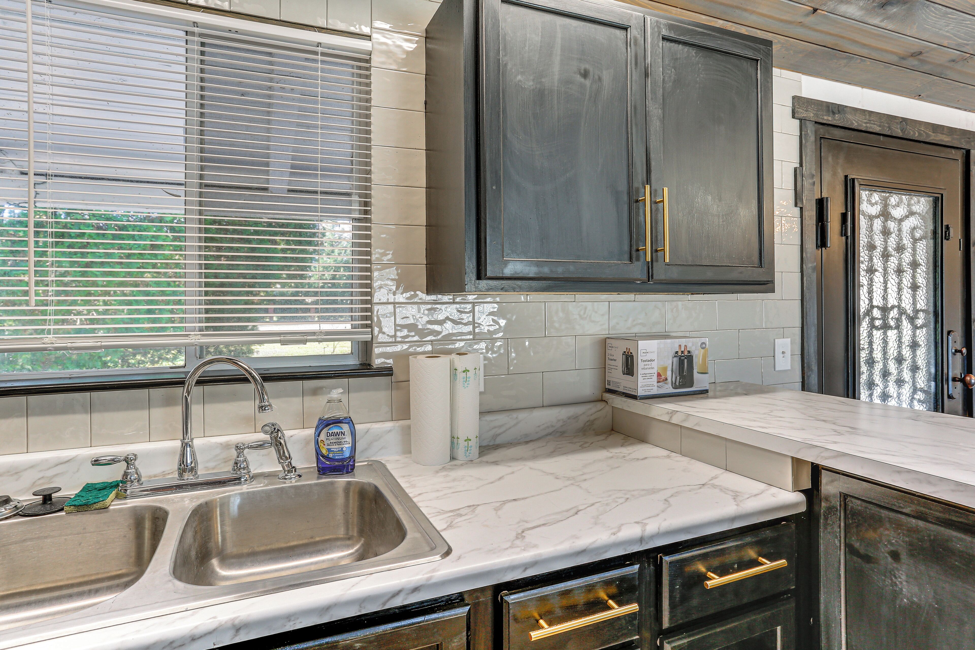 A kitchen with a double sink, stainless steel faucet, and a marble countertop. There are cabinets with gold handles and a window with blinds.