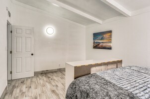Un dormitorio con piso de madera de diseño espiga, una puerta blanca, una mesita de noche, una cama con colcha estampada floral y un cuadro de un atardecer en la pared.