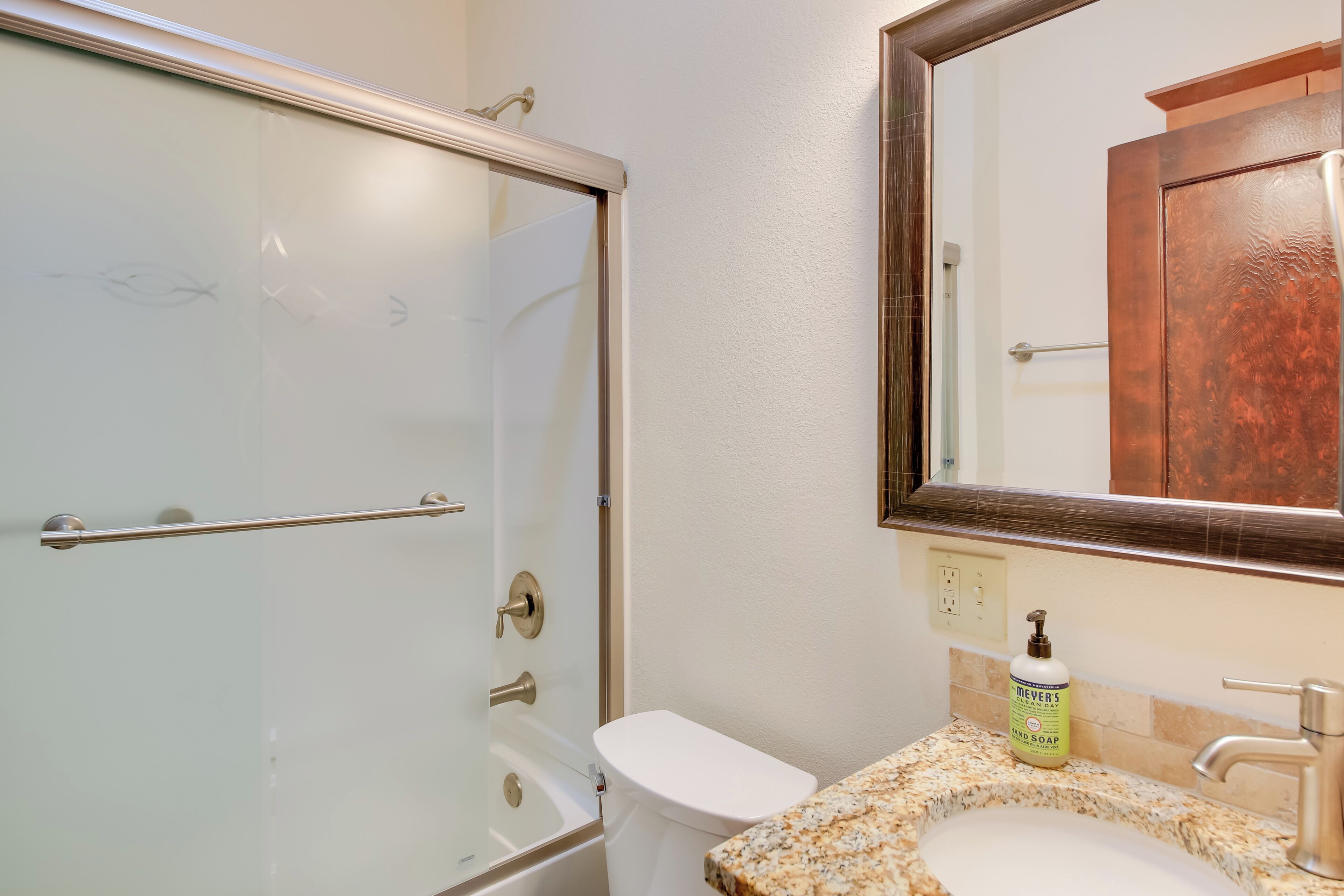 A bathroom with a glass shower door, a white toilet, a granite sink, a mirror, and a bottle of hand soap.