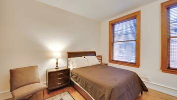 A bedroom with a bed, nightstand, chair, and a window with blinds.
