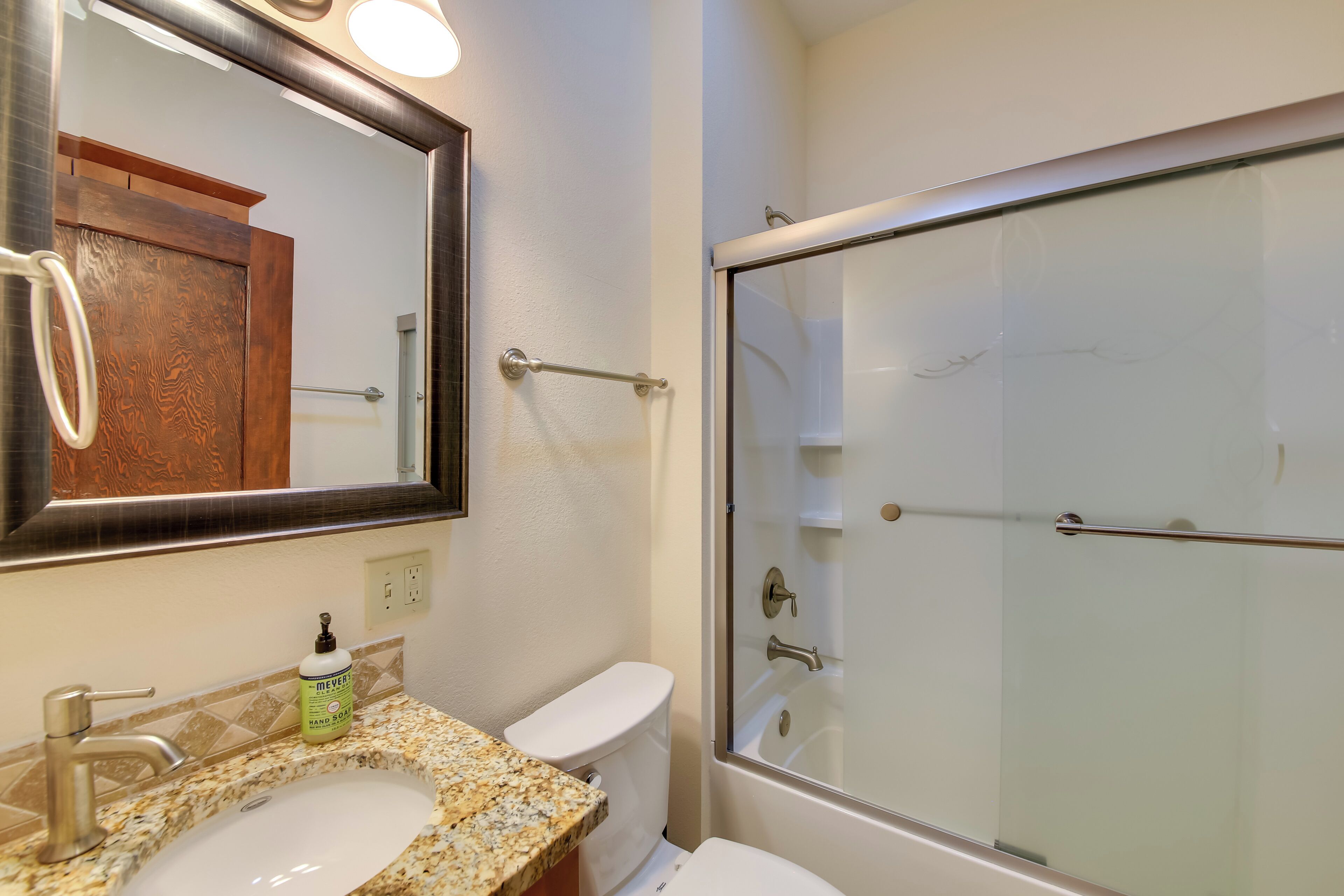 Un baño con lavamanos de encimera de granito, espejo, inodoro y ducha con mampara de vidrio.