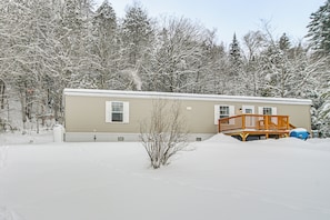 A single-story mobile home with a wooden deck, surrounded by snow-covered trees. - Near Trails & Vast Access! Modern Burke Home Base (East Burke)