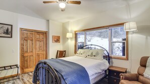 A bedroom with a bed, wooden floor, a ceiling fan, a window with a view of snow outside, a nightstand, and a chair. - Scenic Creekside Retreat! Rocky Mountain Cabin (Dumont)