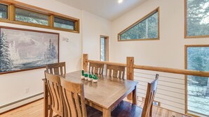 A dining area with a wooden table and chairs, a painting of a snowy landscape, and large windows with a view of trees. - Scenic Creekside Retreat! Rocky Mountain Cabin (Dumont)