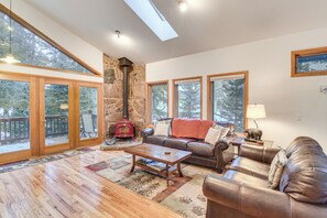 A spacious living room with a stone fireplace, leather sofas, a wooden coffee table, and large windows offering a view of the outdoors. - Scenic Creekside Retreat! Rocky Mountain Cabin (Dumont)